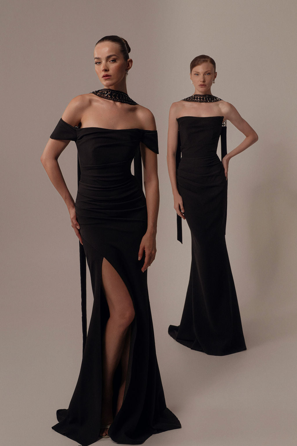 שתי נשים לובשות שמלות אלגנטיות שחורות עם כתפיים חשופות מקולקציית הערב 2026 של דרור קונטנטו, עם חתכים גבוהים וקישוטים בקו הצוואר. האחת עומדת עם ידיים לצדי גופה, והשנייה עם יד על מותנה על רקע אחיד.