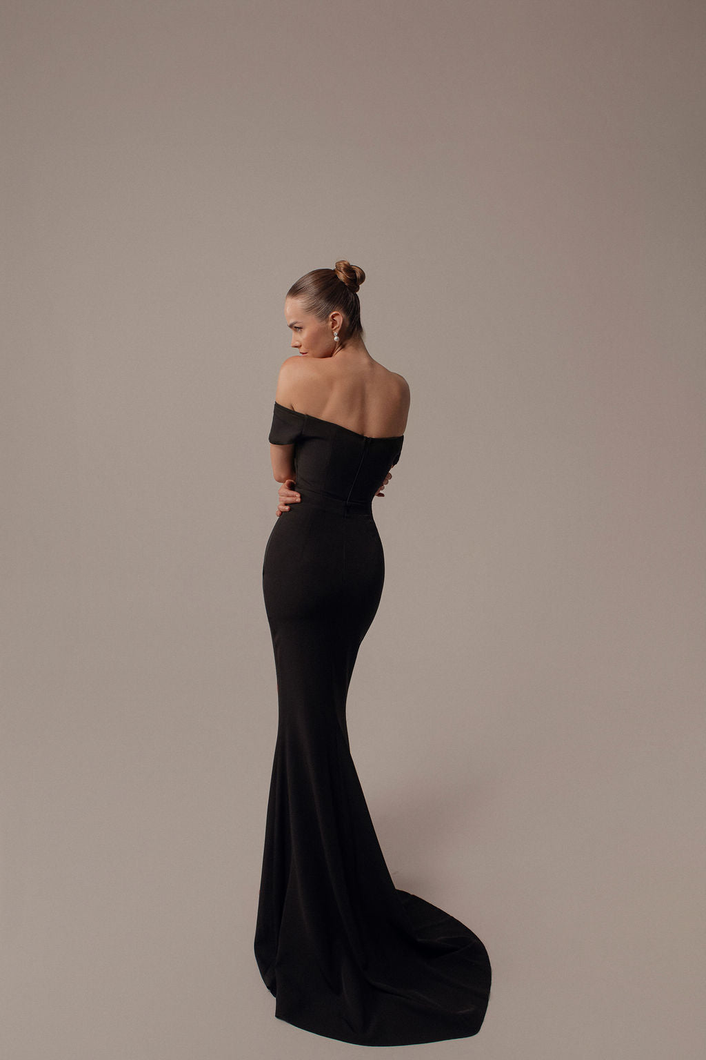 אישה עם שיער אסוף בצמה מפנה את גבה למצלמה, לבושה בשמלת Rona מקולקציית הערב 2026 של דרור קונטנטו — שמלת ערב שחורה עם כתפיים חשופות — עומדת על רקע ניטרלי, זרועותיה שלובות בעדינות.
