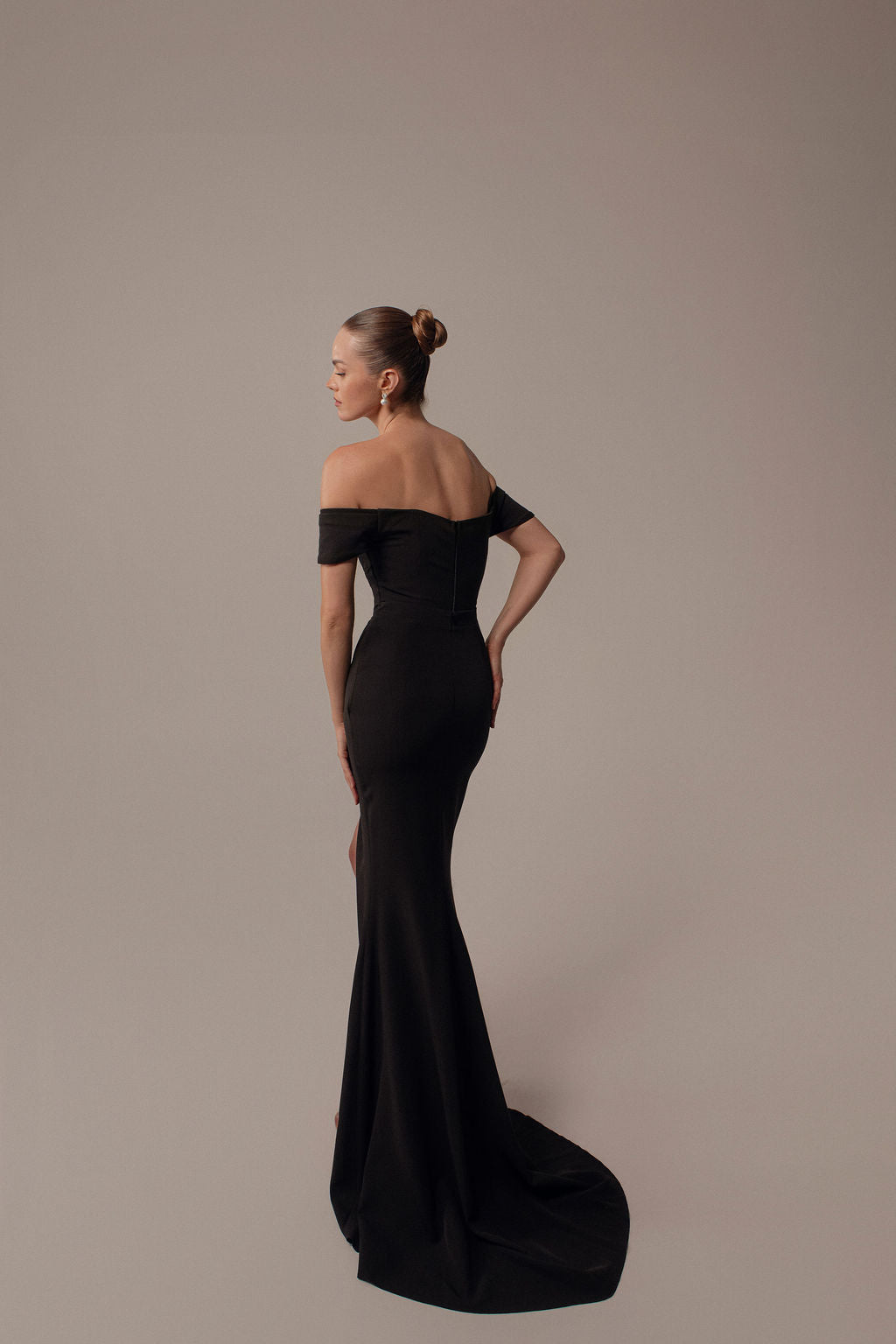 אישה לבושה בשמלת Rona מקולקציית ערב 2026 של דרור קונטנטו — שמלת ערב שחורה באורך מלא עם כתפיים חשופות — עומדת על רקע בז', מביטה מעבר לכתפה, שיערה אסוף בקוקו חלק.