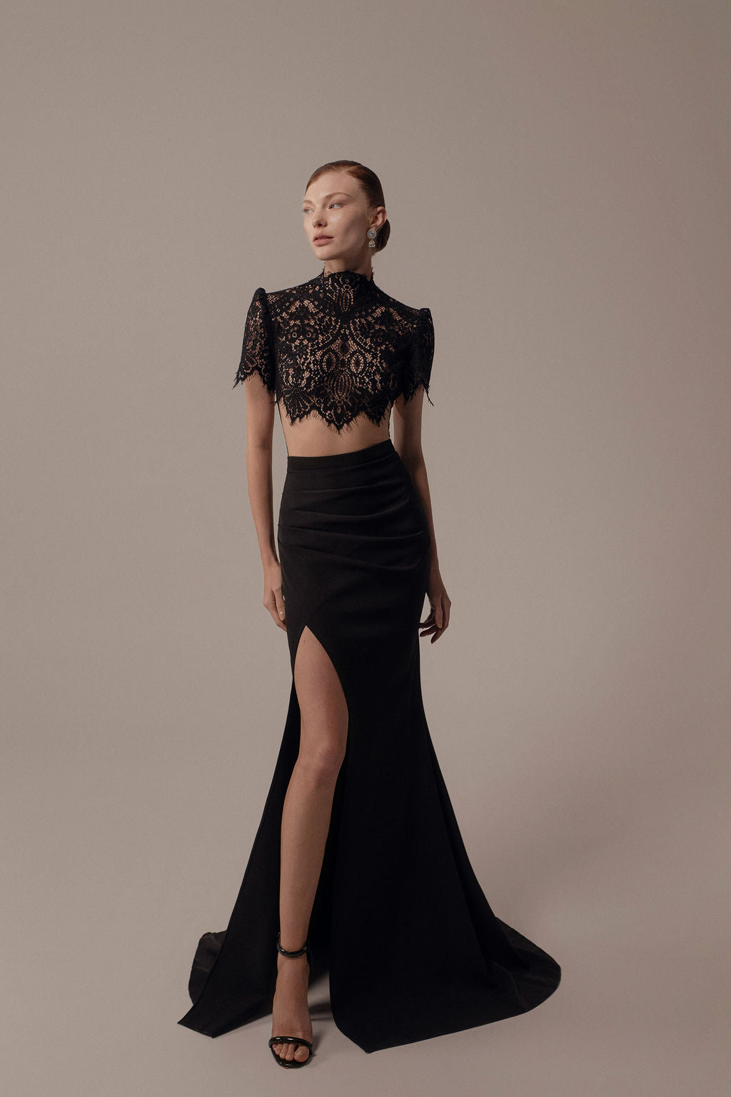 אישה עומדת בסטודיו לבושה בשמלת Hila מקולקציית ערב 2026: חולצת תחרה שחורה קצרה עם שרוולים קצרים וחצאית ארוכה עם חתך גבוה. שיערה אסוף לאחור והיא נועלת סנדלים שחורים עם עקבים גבוהים.