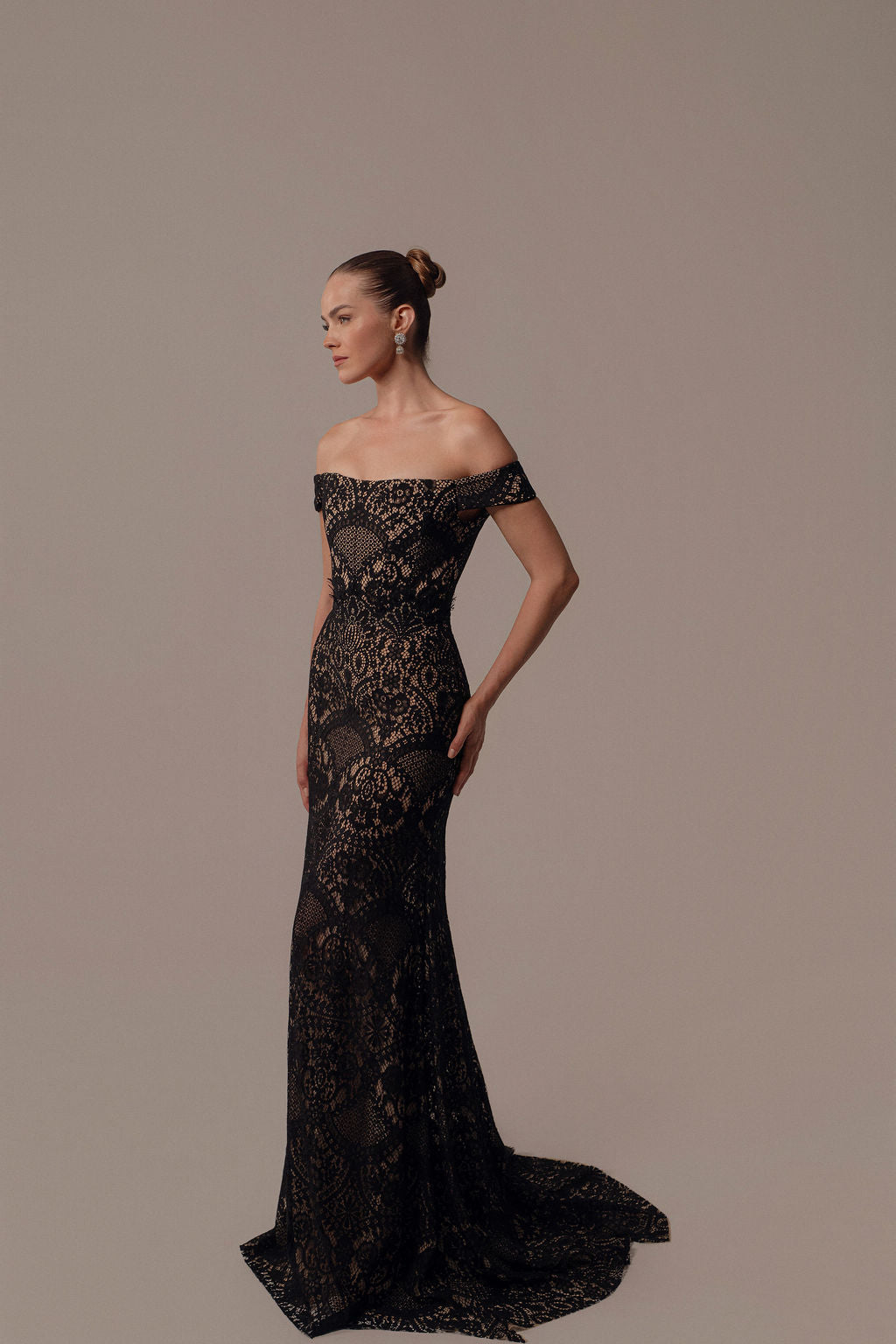 אישה עומדת בפרופיל לבושה בשמלת Neta מקולקציית הערב 2026 של דרור קונטנטו. השמלה האלגנטית עם כתפיים חשופות כוללת תחרה שחורה, שובל ארוך, עגילים בולטים ותסרוקת אסופה ומסודרת על רקע ניטרלי.