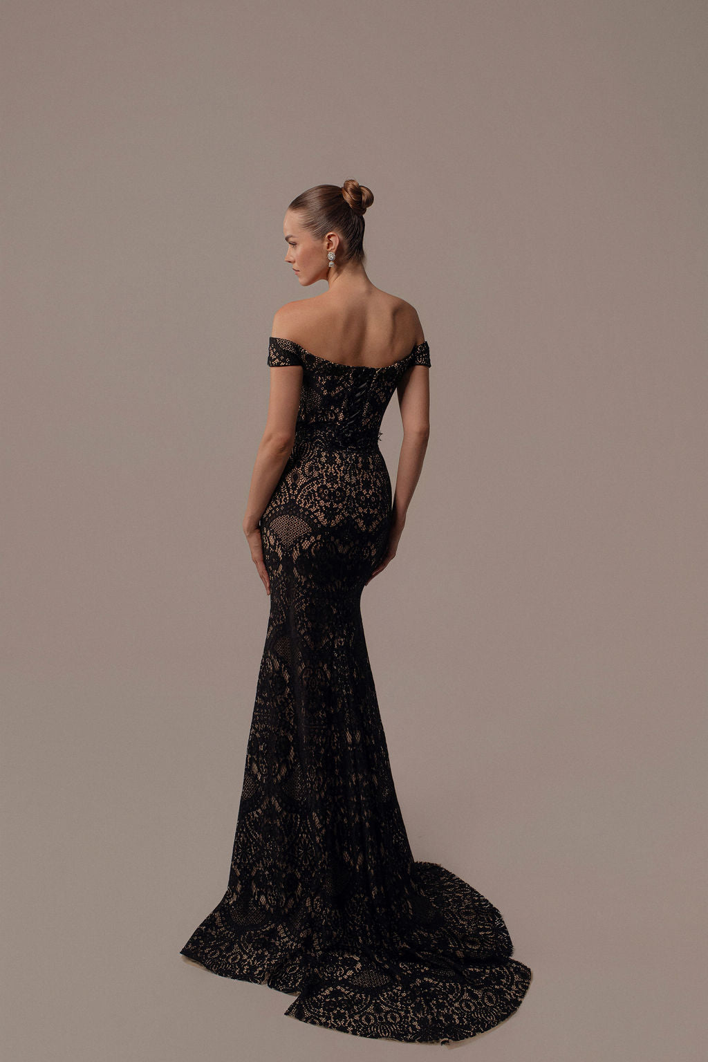 אישה עם שיער אסוף לחבילה לובשת את שמלת Neta מקולקציית ערב 2026, שמלת ערב שחורה באורך הרצפה עם כתפיים חשופות, גזרה צמודה, שובל מתרחב ופרטי תחרה מורכבים, המוצגים כשהיא מפנה את גבה.