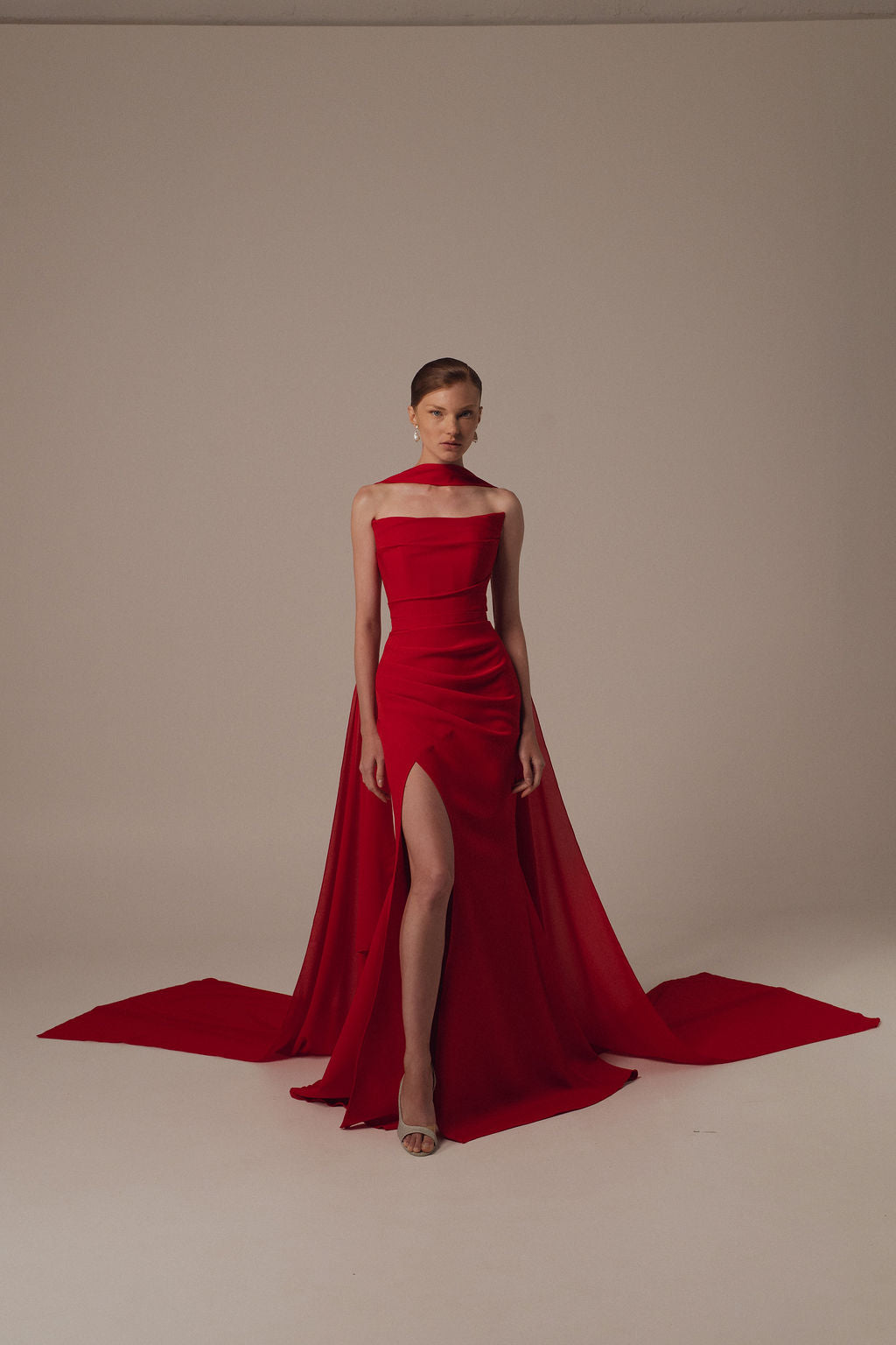 אישה מצטלמת בסטודיו על רקע בז', לבושה בשמלת מירית מקולקציית ערב 2026 — שמלת ערב אדומה ללא כתפיות, באורך מלא, עם חתך גבוה ושובל דרמטי — עם שיער אסוף לאחור ועקבים בגוון ניטרלי.