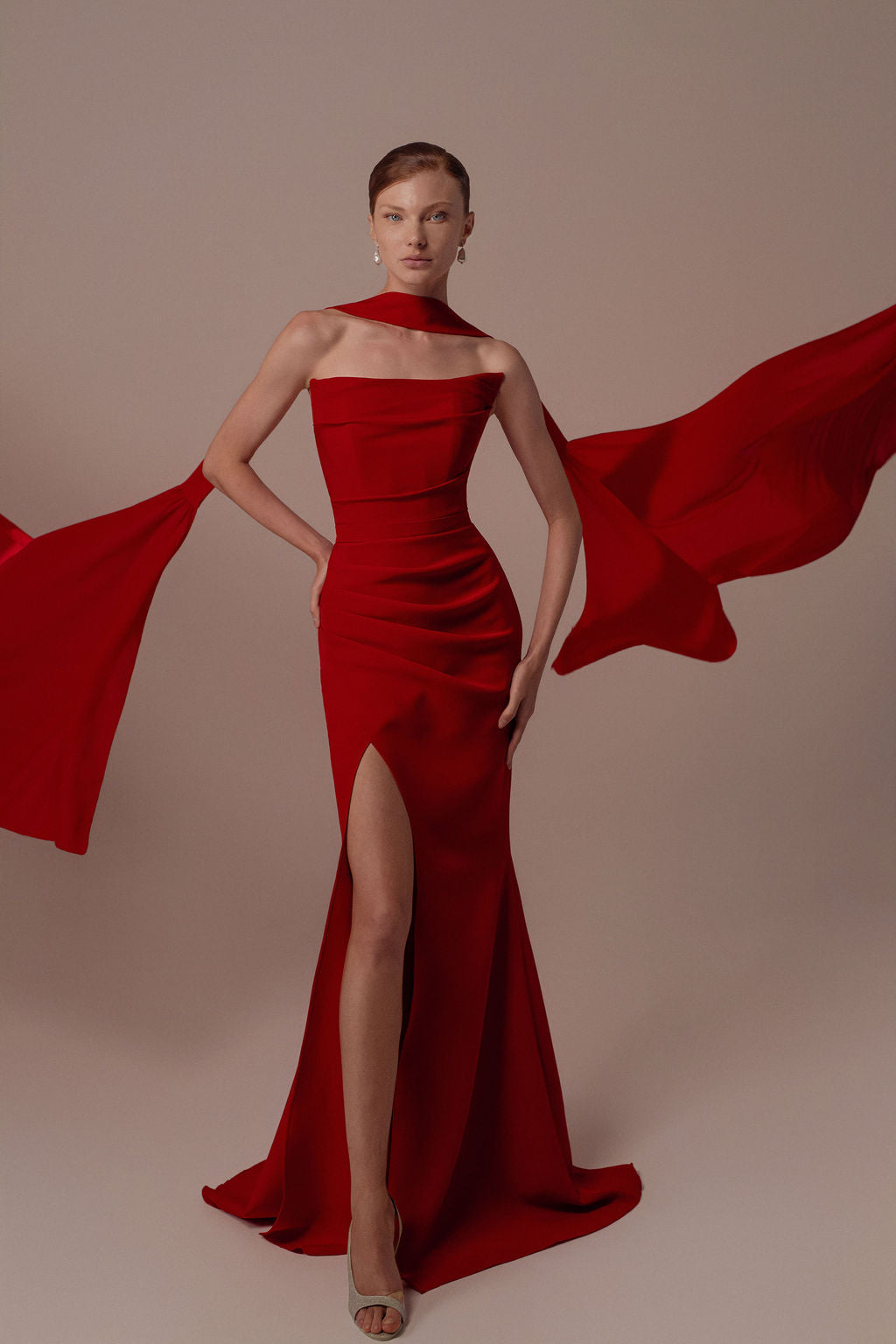 אישה מצטלמת בביטחון בשמלת Tessa מקולקציית ערב 2026, שמלת ערב אדומה ללא כתפיות באורך הרצפה מבית דרור קונטנטו. הבד הדרמטי זורם מזרועותיה ומוסיף נופך דינמי על רקע ניטרלי.