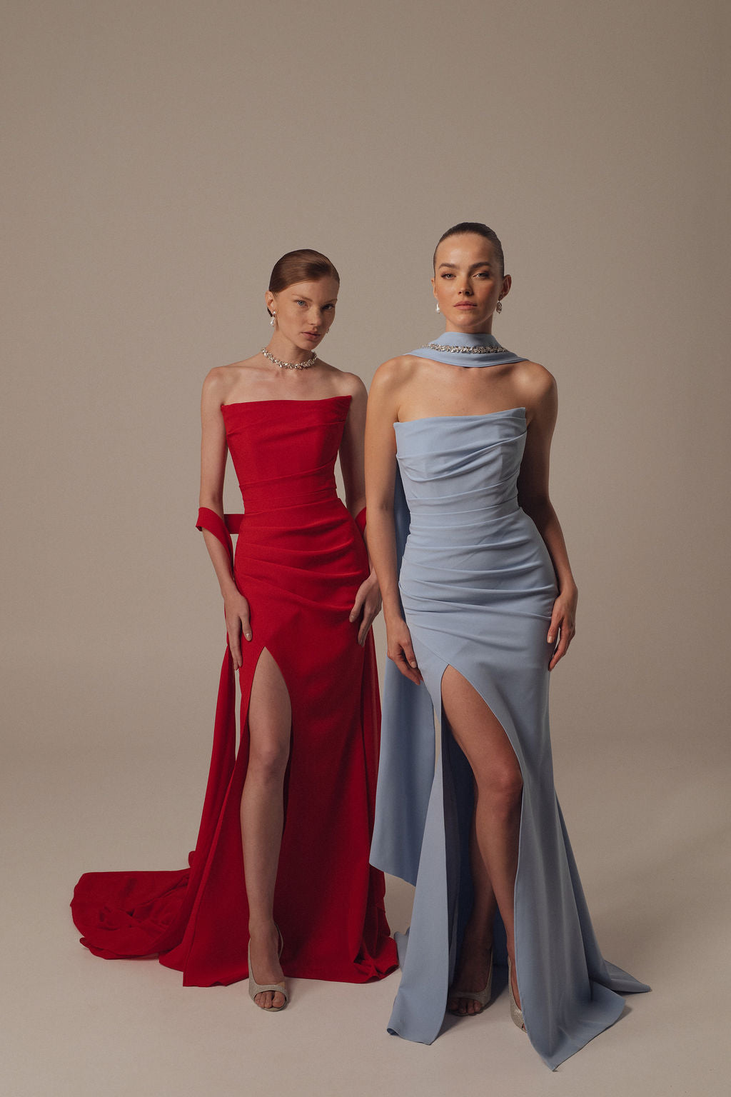 שתי נשים לובשות את שמלת Miri מקולקציית ערב 2026 — האחת בשמלה אדומה בוהקת עם שובל, והשנייה בשמלה כחולה בהירה. שתי השמלות הן ללא כתפיות עם חתכים עד הירך, בשילוב עם שרשראות בולטות ותסרוקות אלגנטיות.