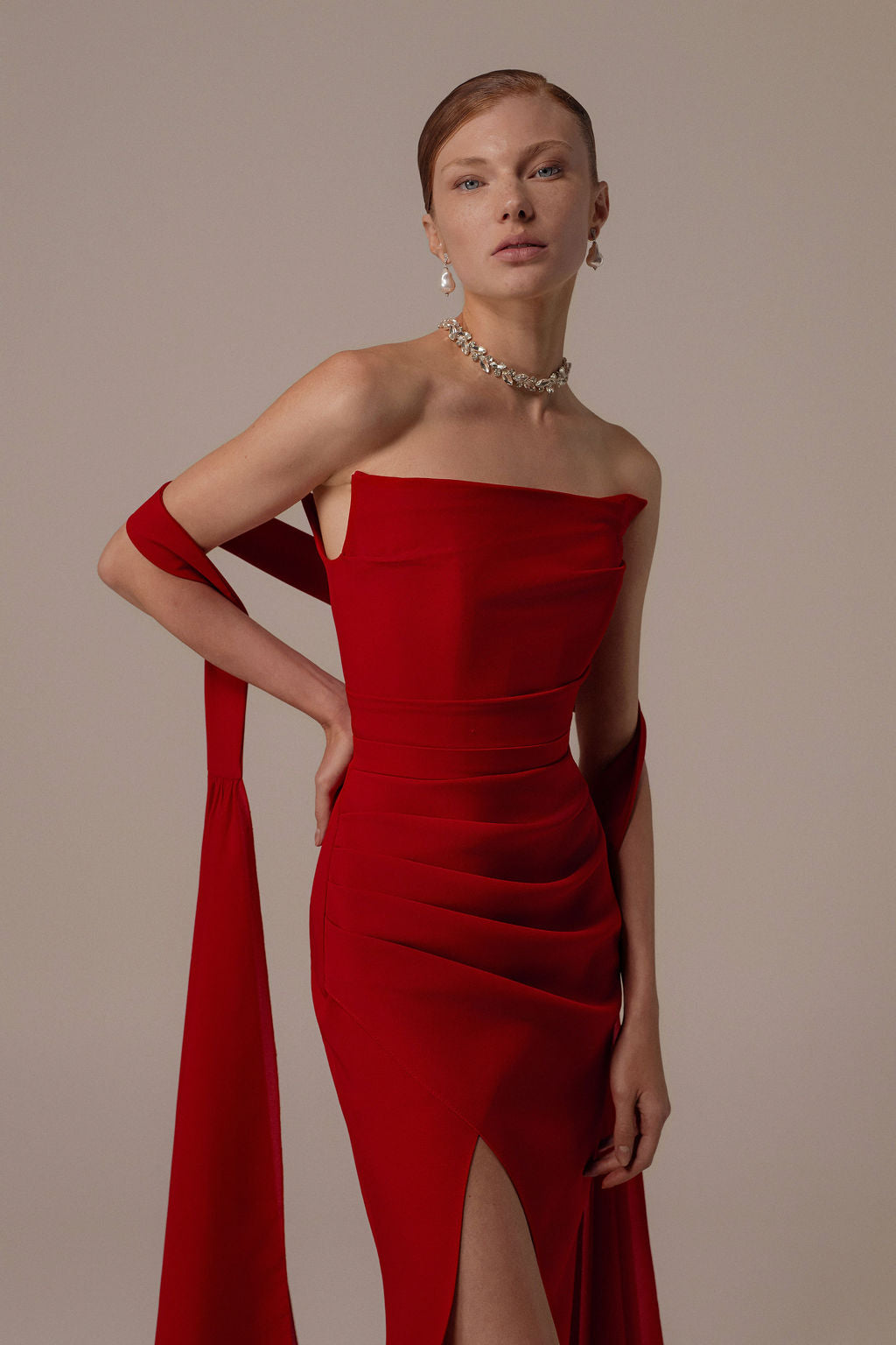 אישה מצטלמת על רקע אחיד בשמלת Tessa מקולקציית Evening 2026 — שמלת ערב אדומה צמודה ללא כתפיות עם בד דראפי וחתך עד הירך — בשילוב עגילי טיפה, שרשרת וצעיף אדום תואם.