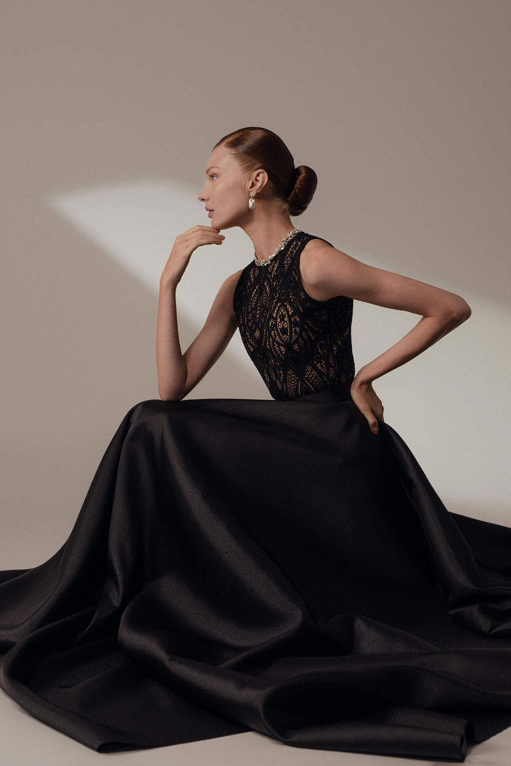 אישה ג'ינג'ית עם תסרוקת אסופה אלגנטית יושבת בחינניות בשמלת Lia מקולקציית Evening 2026, שמלה שחורה ארוכה עם מחוך תחרה. היא פוזלת באלגנטיות, מביטה לצד על רקע ניטרלי.