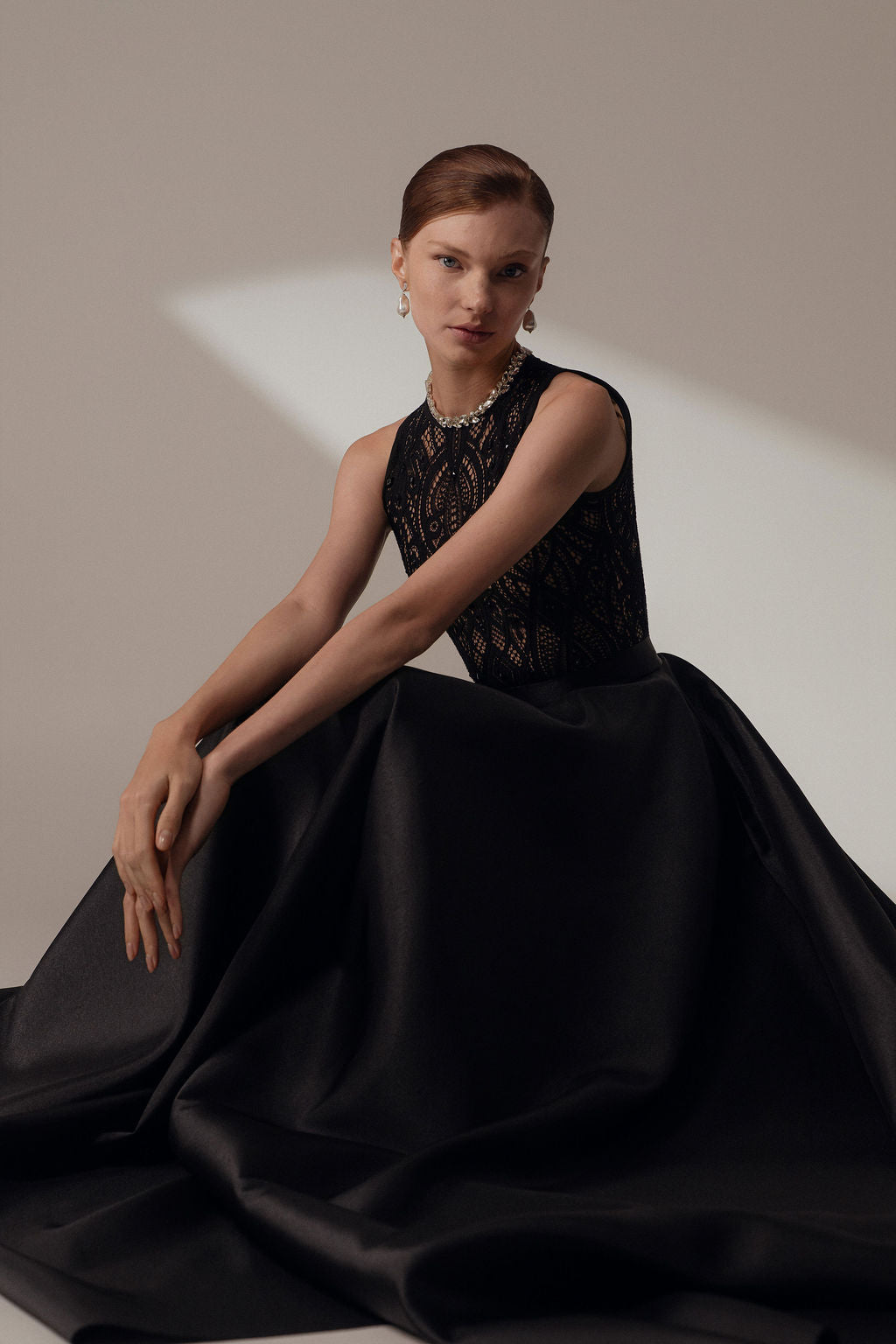 אישה בעלת עור בהיר ושיער אדום פוזלת בחינניות בשמלת Lia מקולקציית הערב 2026 של דרור קונטנטו, הכוללת חולצת תחרה שחורה ללא שרוולים וחצאית נפוחה, וידיה מונחות באלגנטיות על רקע ניטרלי.