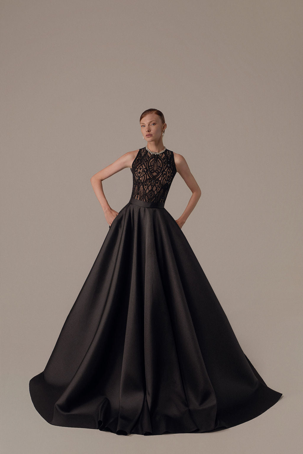 אישה עומדת בביטחון בשמלת Lia מקולקציית ערב 2026 — שמלת ערב שחורה ללא שרוולים באורך הרצפה עם מחוך תחרה שקוף ומורכב, על רקע בהיר וחלק.