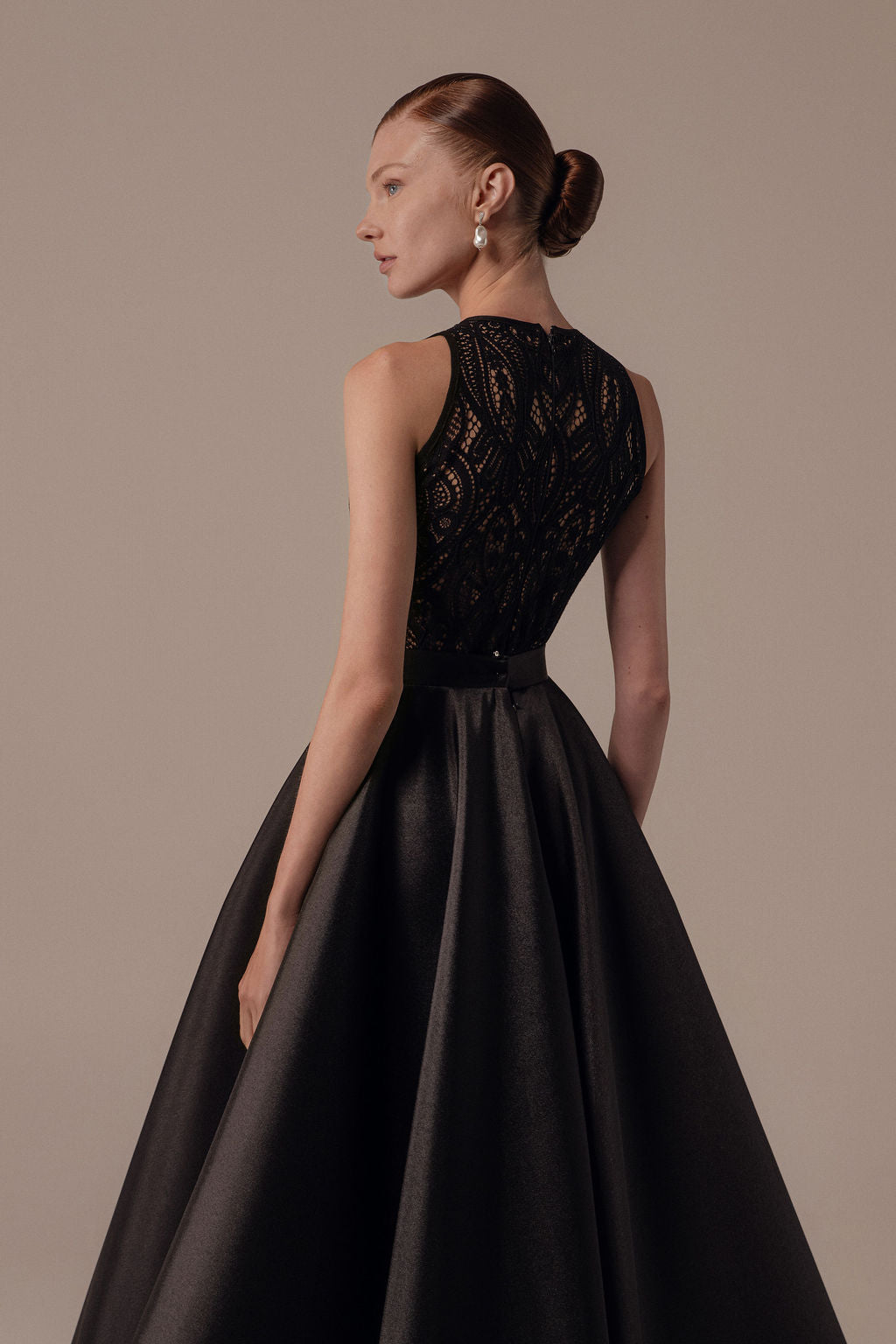 אישה עם שיער חום-אדמדם אסוף בקוקו אלגנטי לובשת את שמלת Lia מקולקציית Evening 2026 — שמלת ערב שחורה ללא שרוולים עם מחוך תחרה וחצאית סאטן מלאה — עומדת על רקע ניטרלי ומביטה לצד.