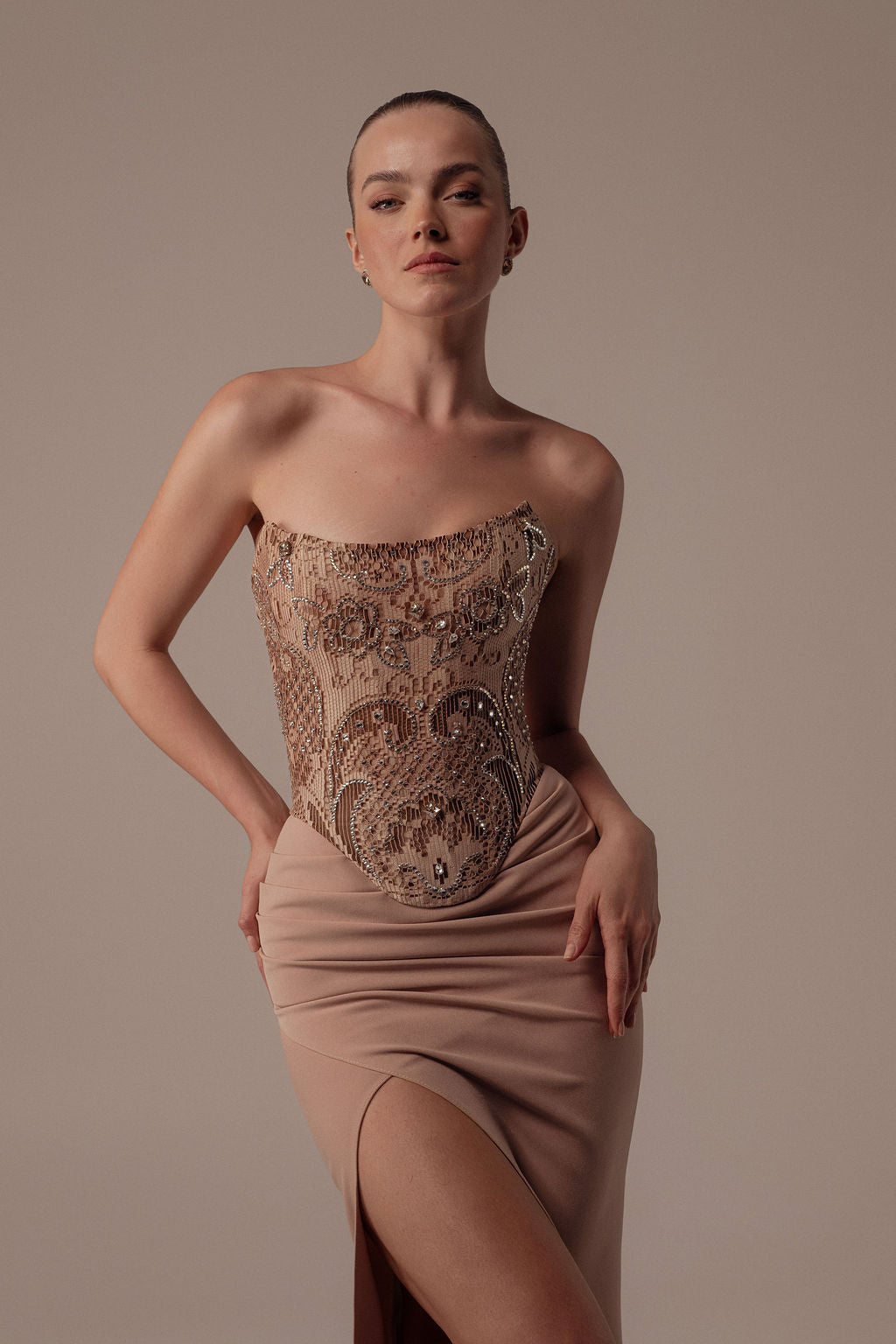 אישה מצטלמת בביטחון בשמלת "Dana" מקולקציית ערב 2026 של דרור קונטנטו — שמלת ערב ללא כתפיות בגוון ניוד עם מחשוף מעוטר בחרוזים וחתך גבוה — על רקע ניטרלי.