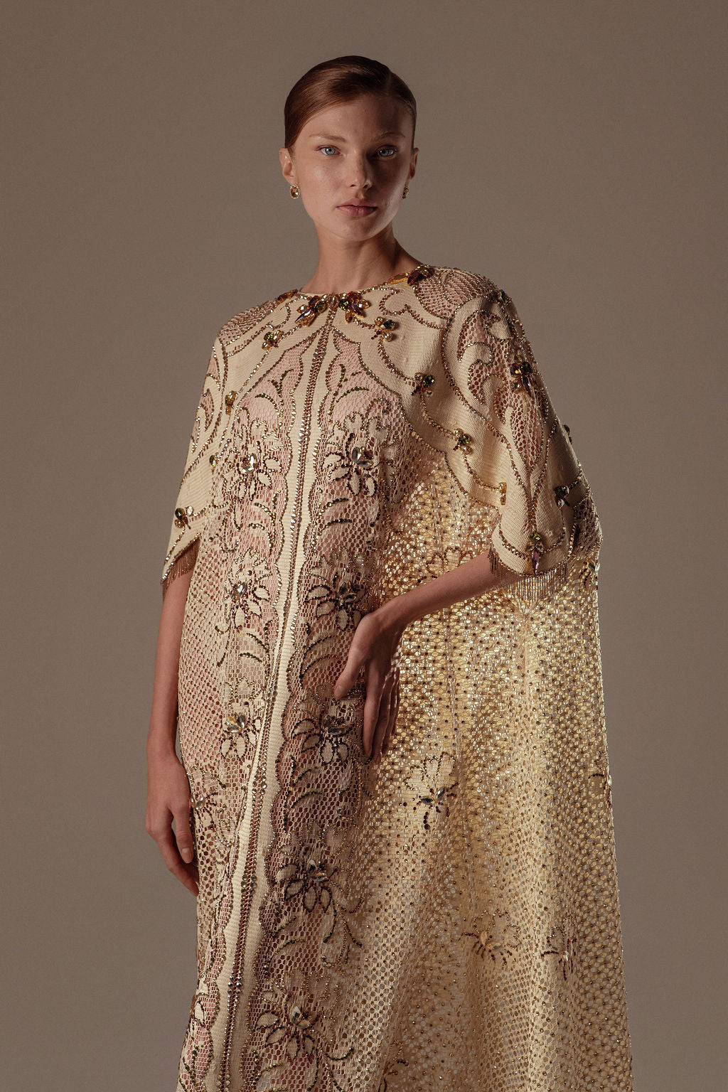 אישה בעלת עור בהיר ושיער חום קצר לובש את שמלת Mira מקולקציית הערב 2026 של דרור קונטנטו — שמלת ערב מעוטרת בצבעי שמנת וזהב עם רקמת פרחים מורכבת — על רקע ניטרלי.