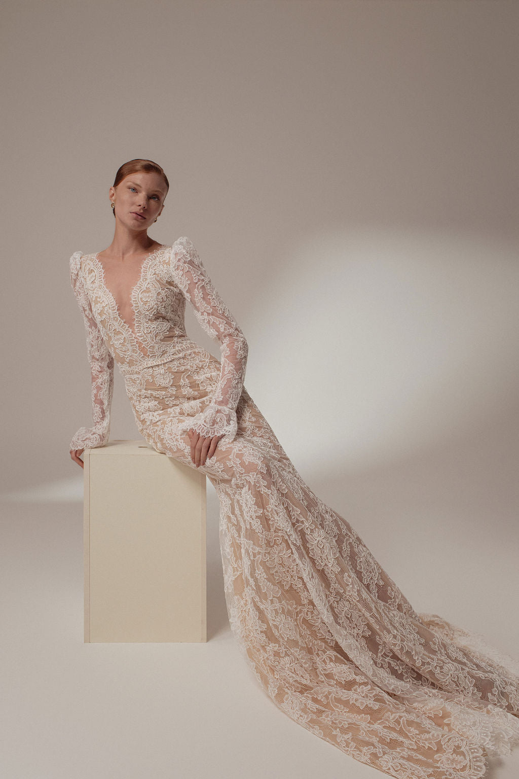 אישה לבושה בשמלת Zara מקולקציית ערב 2026, שמלה ארוכה וצמודה עם תחרה, שרוולים שקופים, מחשוף V עמוק, דוגמת פרחים ושובל דרמטי, יושבת על בלוק שמנת על רקע ניטרלי.