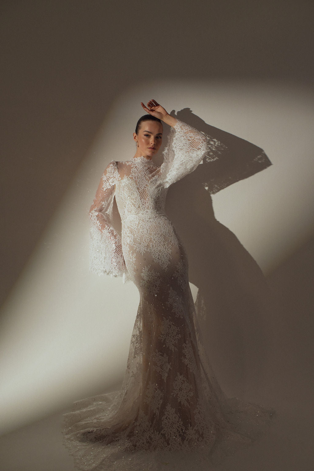 אישה מצטלמת על רקע קיר בשמלת קירה שקופה עם שרוולים ארוכים מקולקציית הערב 2026 של דרור קונטנטו, עם מחוך צמוד ושרוולים מתרחבים, היוצרים צל דרמטי.