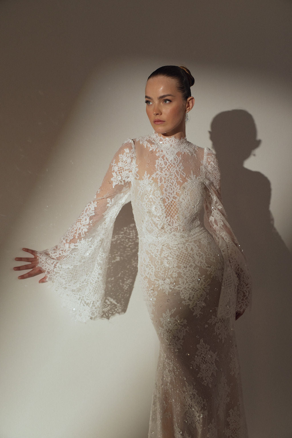 אישה מדגמנת את שמלת Kira מקולקציית ערב 2026 של דרור קונטנטו. האור מטיל צללים מאחוריה, ומדגיש את פרטי התחרה המורכבים ואת השרוולים המתרחבים של השמלה הלבנה השקופה עם השרוולים הארוכים.