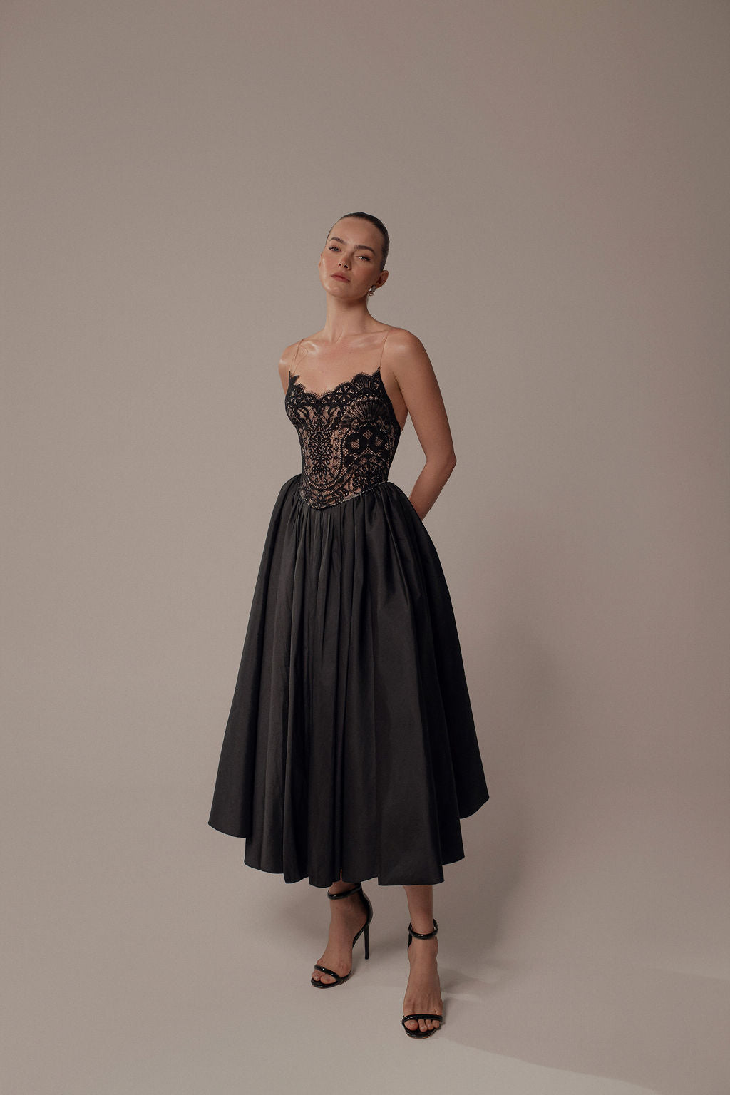 אישה מצטלמת על רקע אחיד בשמלת Luna מקולקציית Evening 2026, עם מחוך שחור צמוד מתחרה וחצאית מלאה. היא עומדת עם ידיים מאחורי הגב, לובשת סנדלים שחורים עם עקבים גבוהים.