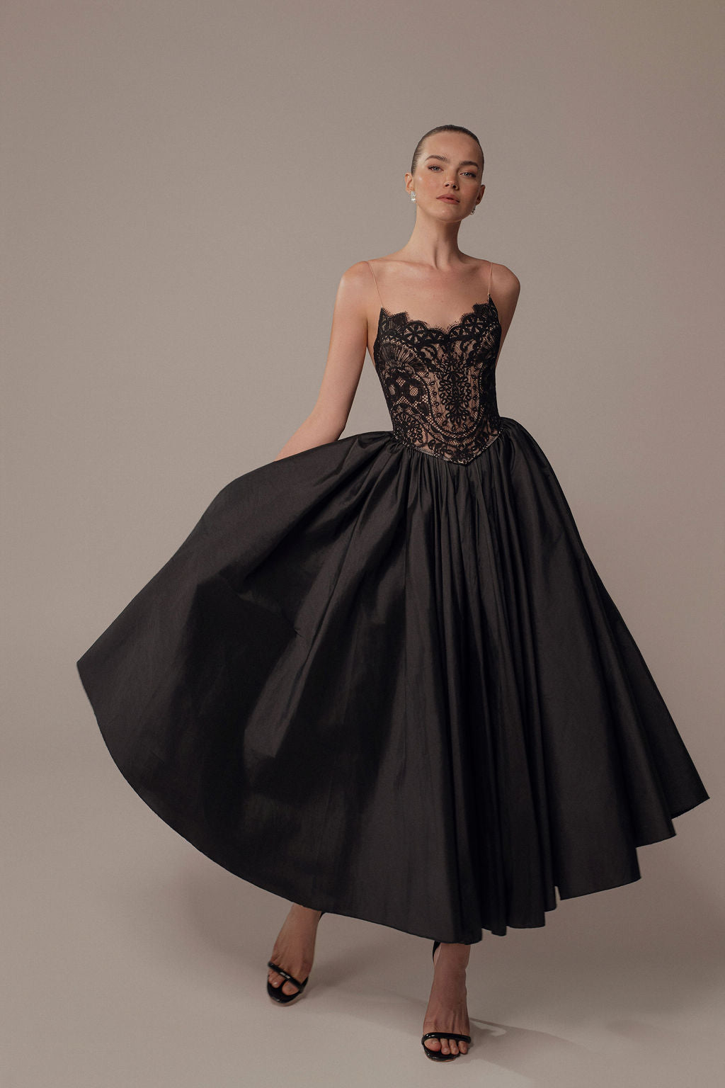 אישה מדגמנת את שמלת Luna מקולקציית ערב 2026 של דרור קונטנטו, עם מחוך תחרה וחצאית מלאה, על רקע בז' חלק, כשהיא נועלת נעלי עקב שחורות.