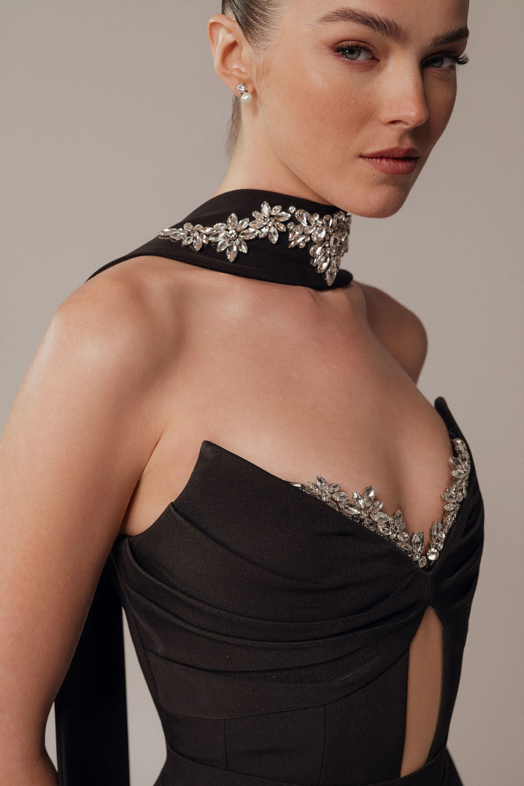 אישה לובשת את שמלת Maya מקולקציית הערב 2026 של דרור קונטנטו — שחורה, ללא כתפיות, עם עיטורי פרחים מריינסטון וצעיף משובץ אבנים — שיערה אסוף לאחור והיא פונה מעט לצד.