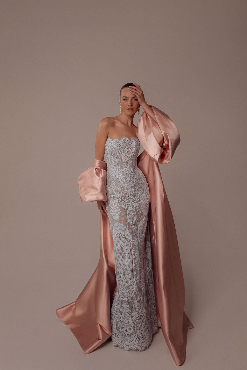 אישה מצטלמת על רקע ניטרלי בשמלת Elia מקולקציית הערב 2026 של דרור קונטנטו — שמלת ערב מחויטת ללא כתפיות, עם תחרה בעלת דוגמאות מורכבות, בשילוב חלוק סאטן ארוך בצבע ורוד סומק.