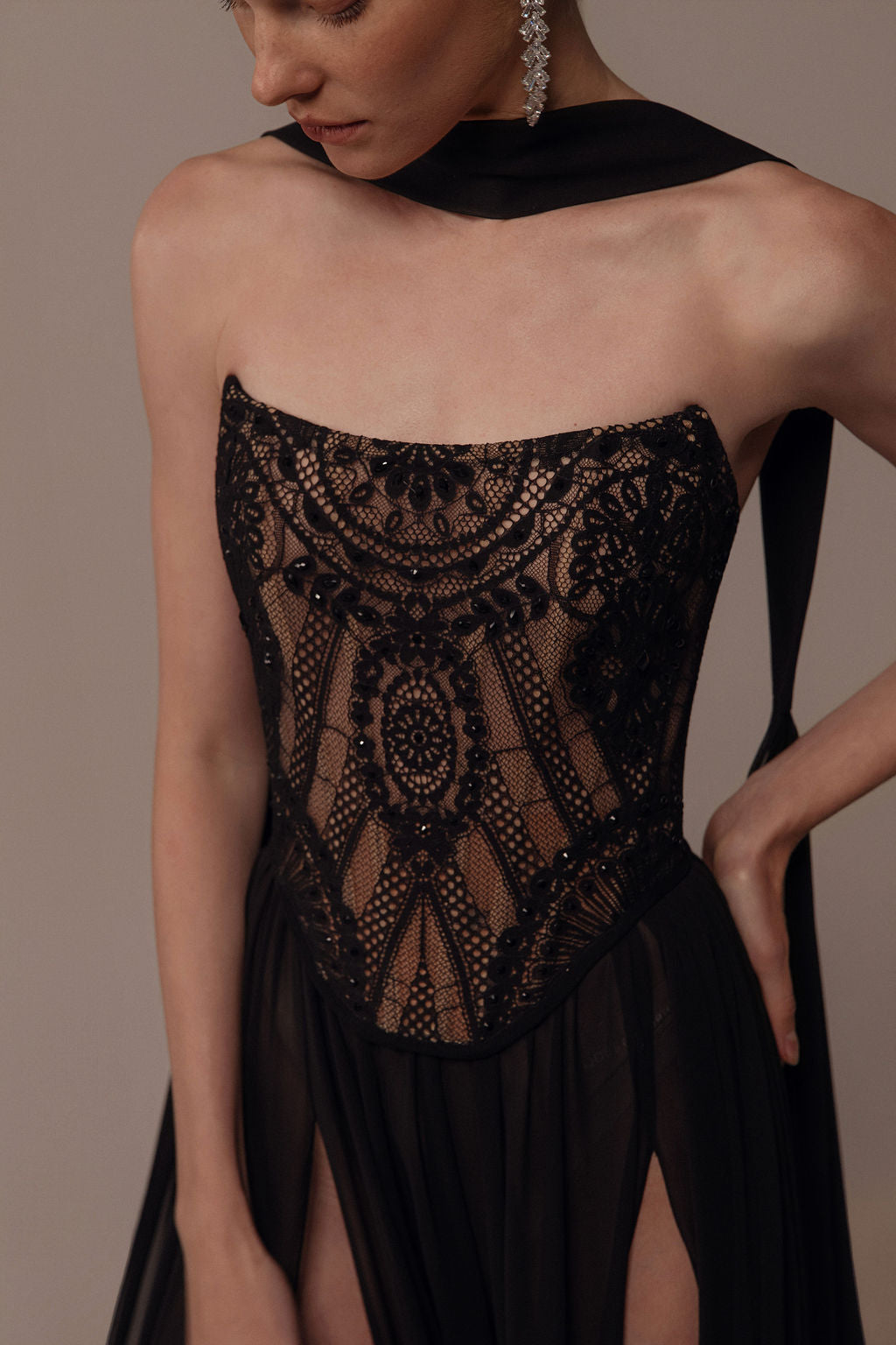 אישה לובשת את שמלת Alma מקולקציית הערב 2026 של דרור קונטנטו — שמלת מחוך שחורה ללא כתפיות עם תחרה בעלת דוגמאות מורכבות וחצאית מתנפנפת. צעיף שחור עוטף את צווארה, והיא עונדת עגילים תלויים.