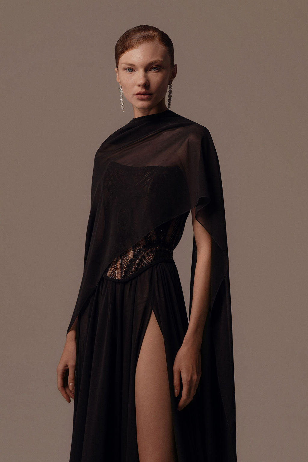 אישה לובשת את שמלת Alma מקולקציית ערב 2026 של דרור קונטנטו — שמלת ערב שחורה שקופה וא-סימטרית עם פרטים מתחרה וחתך גבוה — בשילוב עם שיער אסוף לאחור ועגילי תליון ליצירת מראה אלגנטי של שמלת ערב.