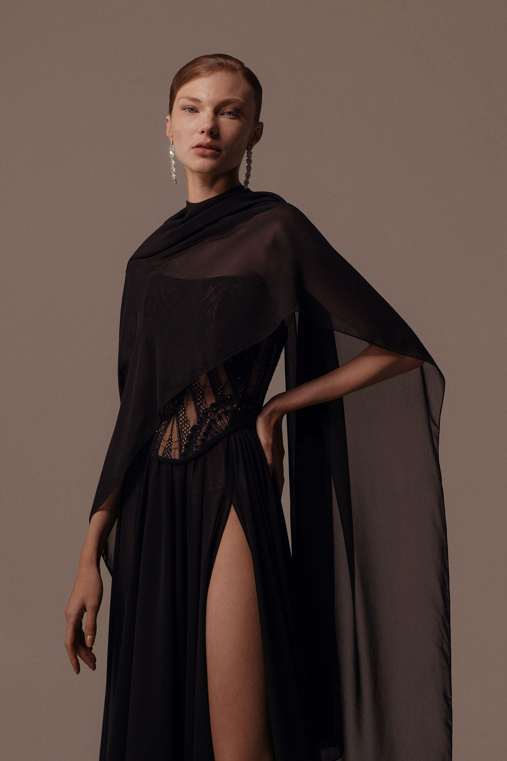 אישה מדגמנת את שמלת "אלמה" מקולקציית ערב 2026 של דרור קונטנטו, העשויה מבד שחור שקוף, עם חתך גבוה, פרטים מתחרה וגילימה דרפית. היא משלימה את המראה עם עגילי טיפה ושיער אסוף לאחור על רקע בז'.