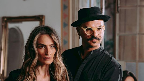 אישה עם שיער ארוך וגלי והבעה רצינית עומדת ליד גבר עם משקפיים, כובע ושפם מסולסל, שניהם בתוך מבנה עם מראה וחלונות מטושטשים ברקע.