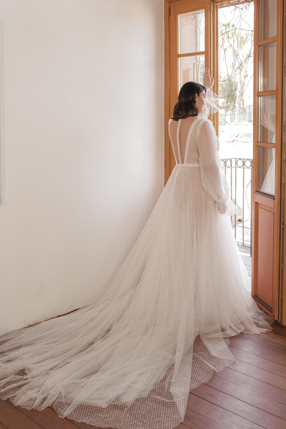 אישה בשמלת כלה ארוכה וזורמת מקולקציית Bridal Plus 2022 - Livia עם שרוולים שקופים וגב פתוח עומדת ליד דלתות כפולות פתוחות, ואור השמש מאיר את השובל האלגנטי.