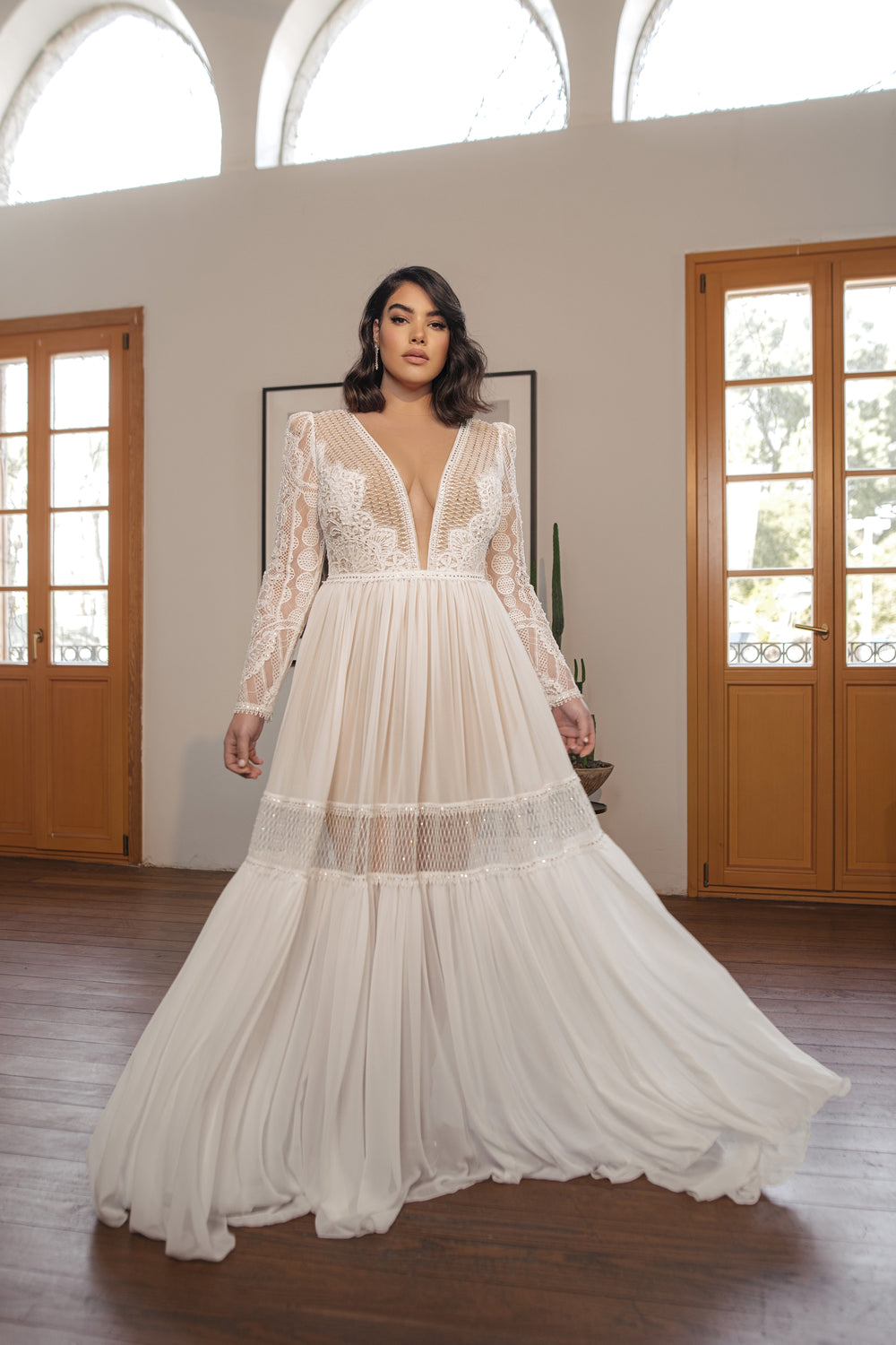 אישה מדגמנת את קולקציית Bridal Plus 2022 - Florence: שמלת תחרה עם שרוולים ארוכים, מחשוף V עמוק וקישוטים, וחצאית מתנפנפת, עומדת בתוך הבית ליד דלתות צרפתיות וחלונות מקושתים - אידיאלית למראה חתונה בוהו שיק.