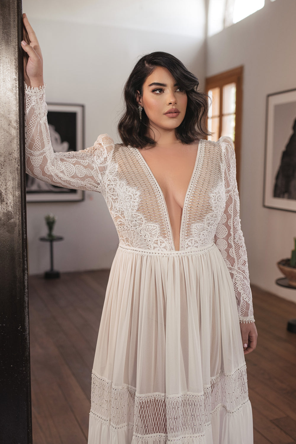 אישה עם שיער כהה וגלי לובשת את שמלת Bridal Plus Collection 2022 - Florence, בעיצוב בוהו שיק עם גימור תחרה ומחשוף V עמוק, עומדת בתוך הבית כשאור רך מסונן מבעד לחלון מאחוריה.