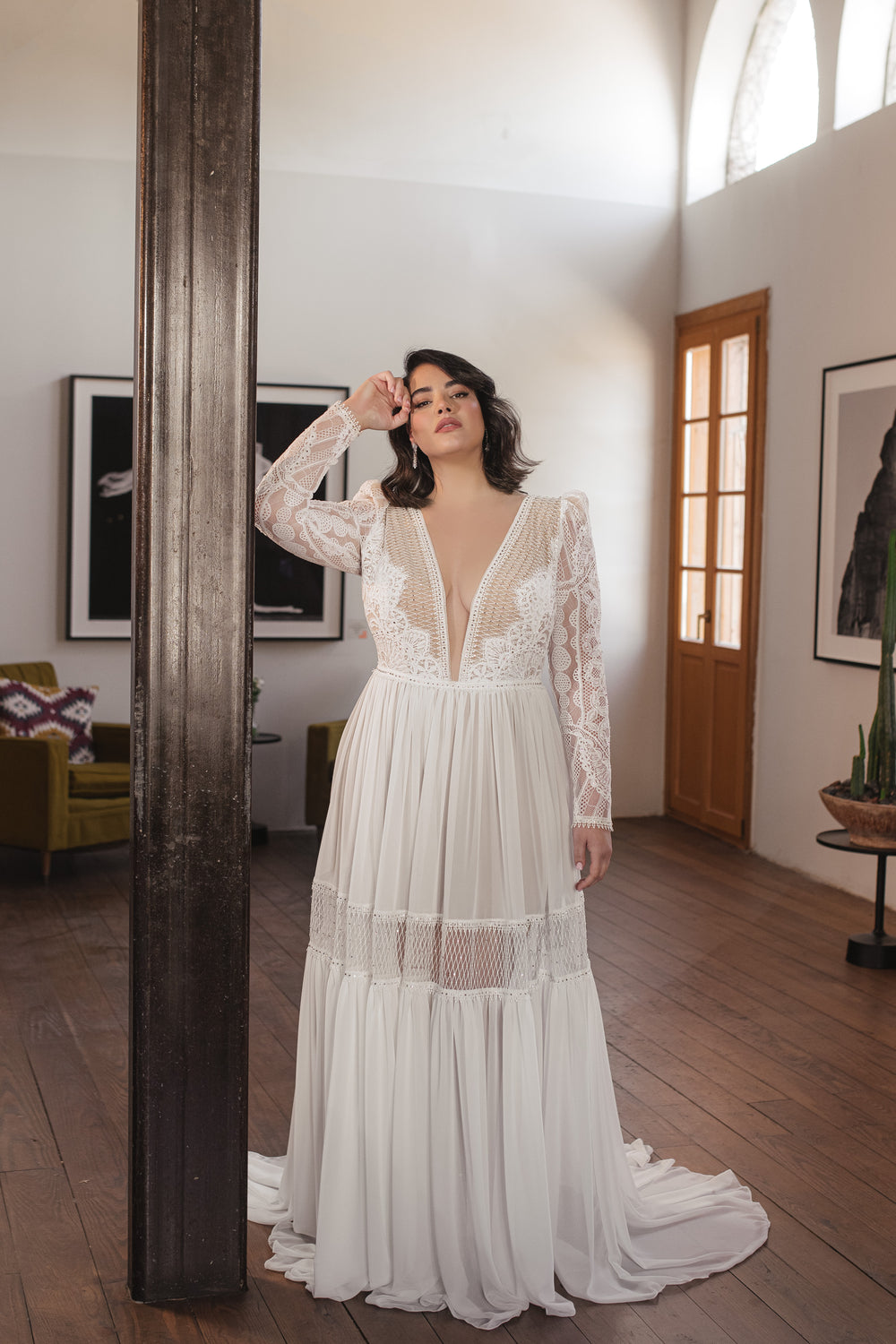 אישה מקולקציית Bridal Plus 2022 - שמלת כלה עם שרוולים ארוכים מתחרה משובצת אבני חן מדגם Florence עומדת בתוך הבית ליד עמוד גבוה וכהה, אור השמש חודר מבעד לחלונות בחדר מודרני עם תמונות ממוסגרות ורצפות עץ.