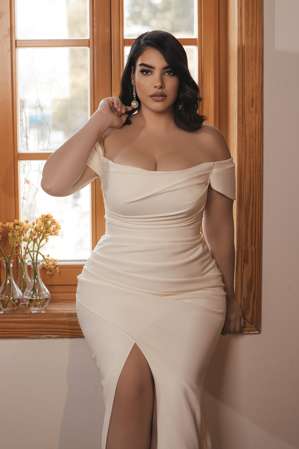 אישה בעלת שיער כהה מקולקציית Bridal Plus 2022 - Aria P by דרור קונטנטו, לבושה בשמלת קרם עם כתפיים חשופות וחתך עד הירך, יושבת על אדן החלון ליד פרחים צהובים, ומביטה בביטחון למצלמה.