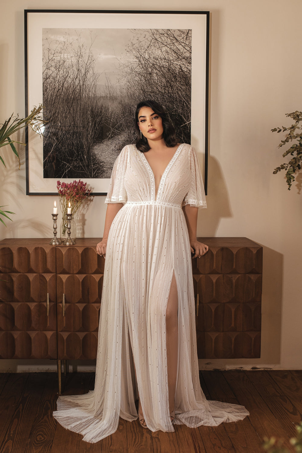 אישה בשמלת כלה לבנה, זורמת, עם מחשוף V, "Bridal Plus Collection 2022 - Jenesis" מאת דרור קונטנטו, עומדת לפני מזנון עץ מתחת לתצלום טבע בשחור-לבן, כששתי נרות דולקים וצמחים משלימים את התמונה.
