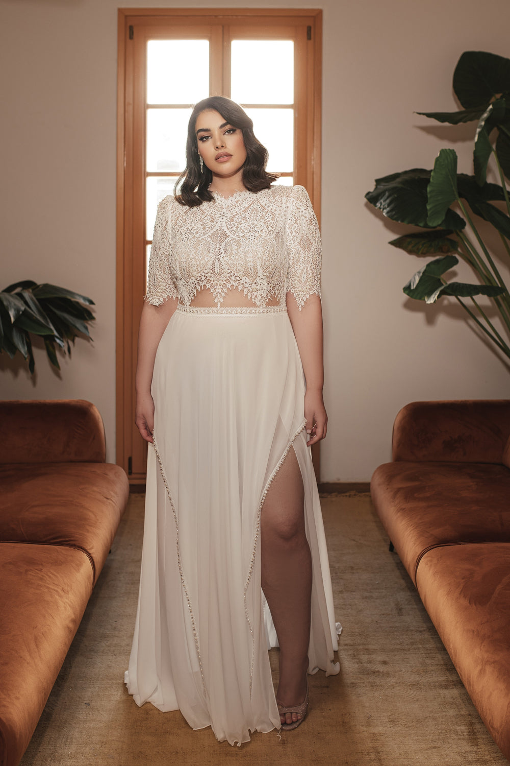 אישה מקולקציית Bridal Plus 2022 - שמלת פנינה עם תחרה משובצת אבני חן וחצאית שיפון עומדת בין שתי ספות חומות בחדר מואר באור רך עם חלון גדול ועציצים.