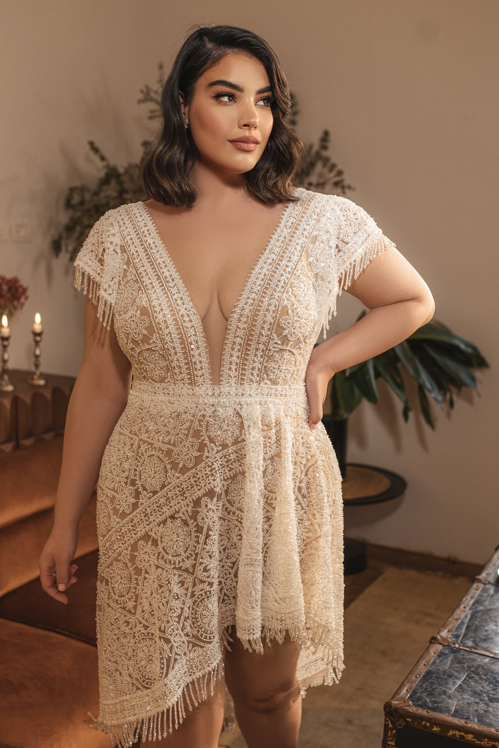 אישה עם שיער כהה וגלי לובשת את קולקציית Bridal Plus 2022 - Giana, שמלת מיני תחרה בצבע שמנת עם מחשוף V עמוק. היא עומדת בתוך הבית ליד ספה חומה, נרות וצמחים - השראה מושלמת למראה כלות במידות גדולות.