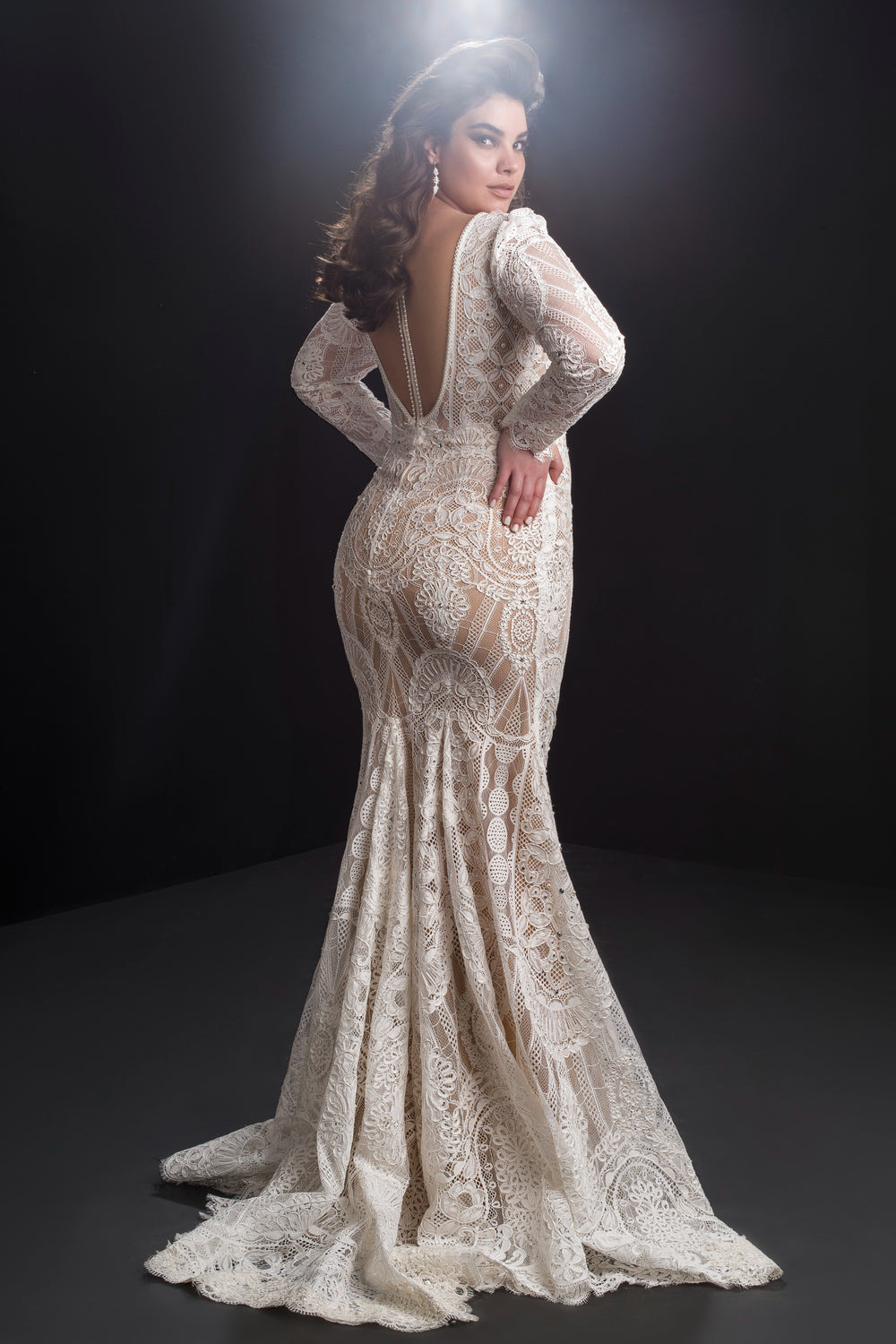אישה מצטלמת בביטחון בשמלת "Bridal Plus Collection 2019 - Rebeca", שמלת כלה מחויטת מתחרה עם שרוולים ארוכים, גב פתוח ושובל דרמטי, על רקע כהה עם תאורה רכה מעליה.