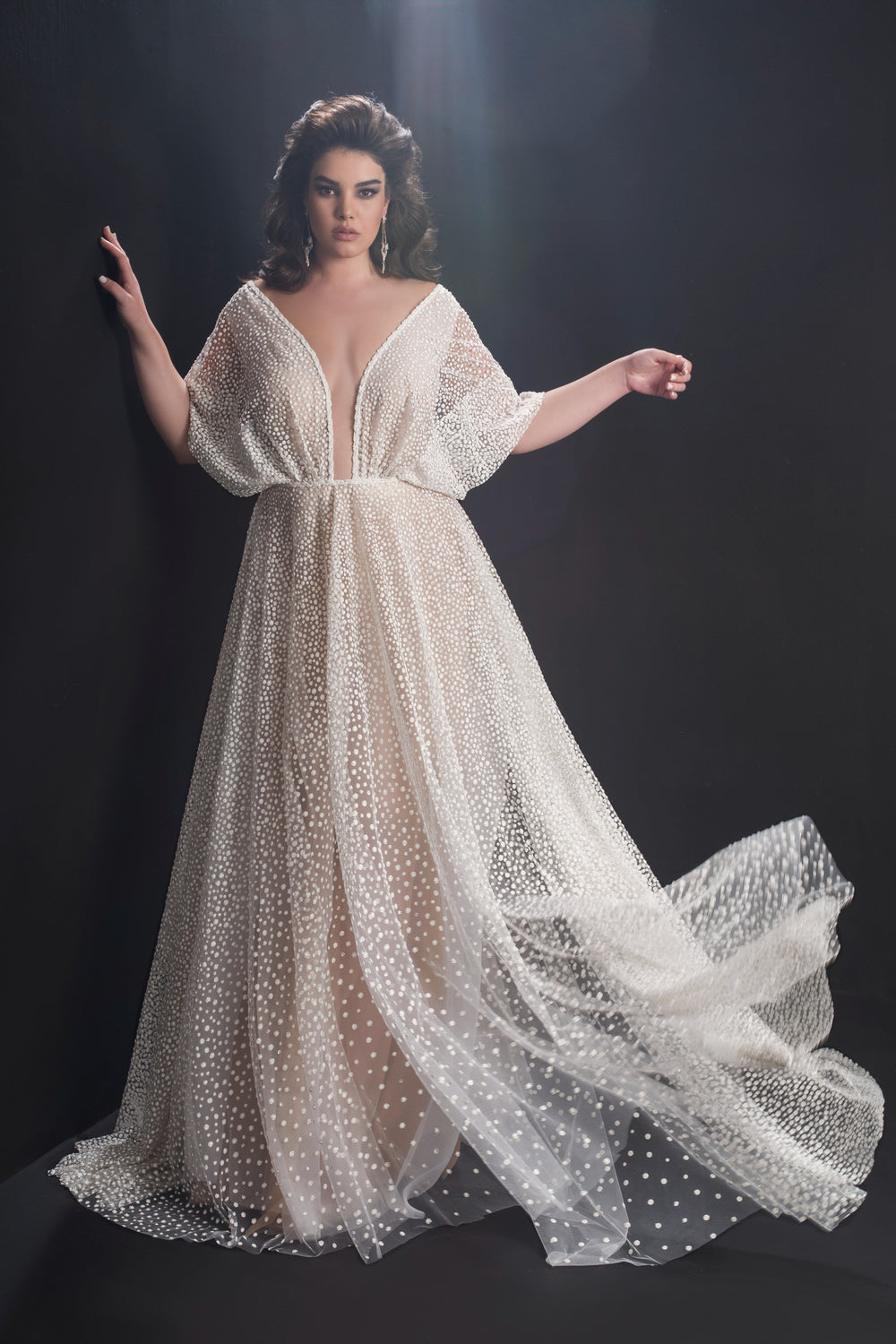 אישה מצטלמת על רקע כהה בשמלת Ellison מקולקציית Bridal Plus 2019, עם בד שקוף ומנוקד, שרוולים רחבים ומחשוף V עמוק. השמלה הלבנה והזורמת מתנפנפת באלגנטיות כשהיא מביטה בביטחון למצלמה.
