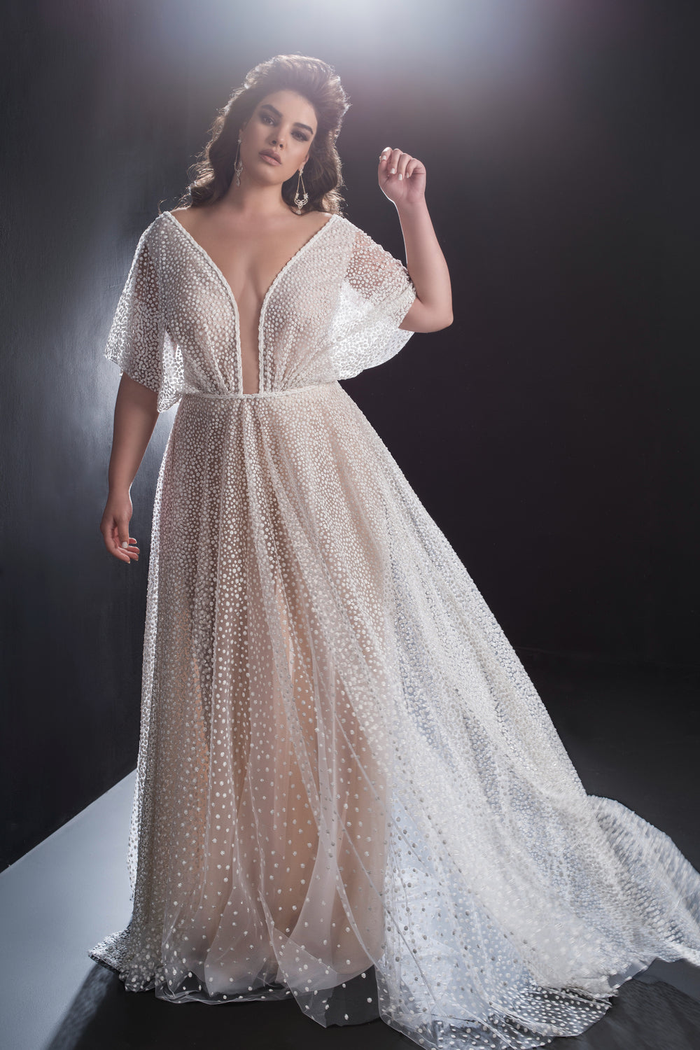 אישה מדגמנת בשמלת אליסון מקולקציית Bridal Plus 2019 — שמלה לבנה שקופה באורך הרצפה עם נקודות, מחשוף V עמוק, שרוולים מתנפנפים ושובל ארוך — על רקע כהה עם תאורה רכה.