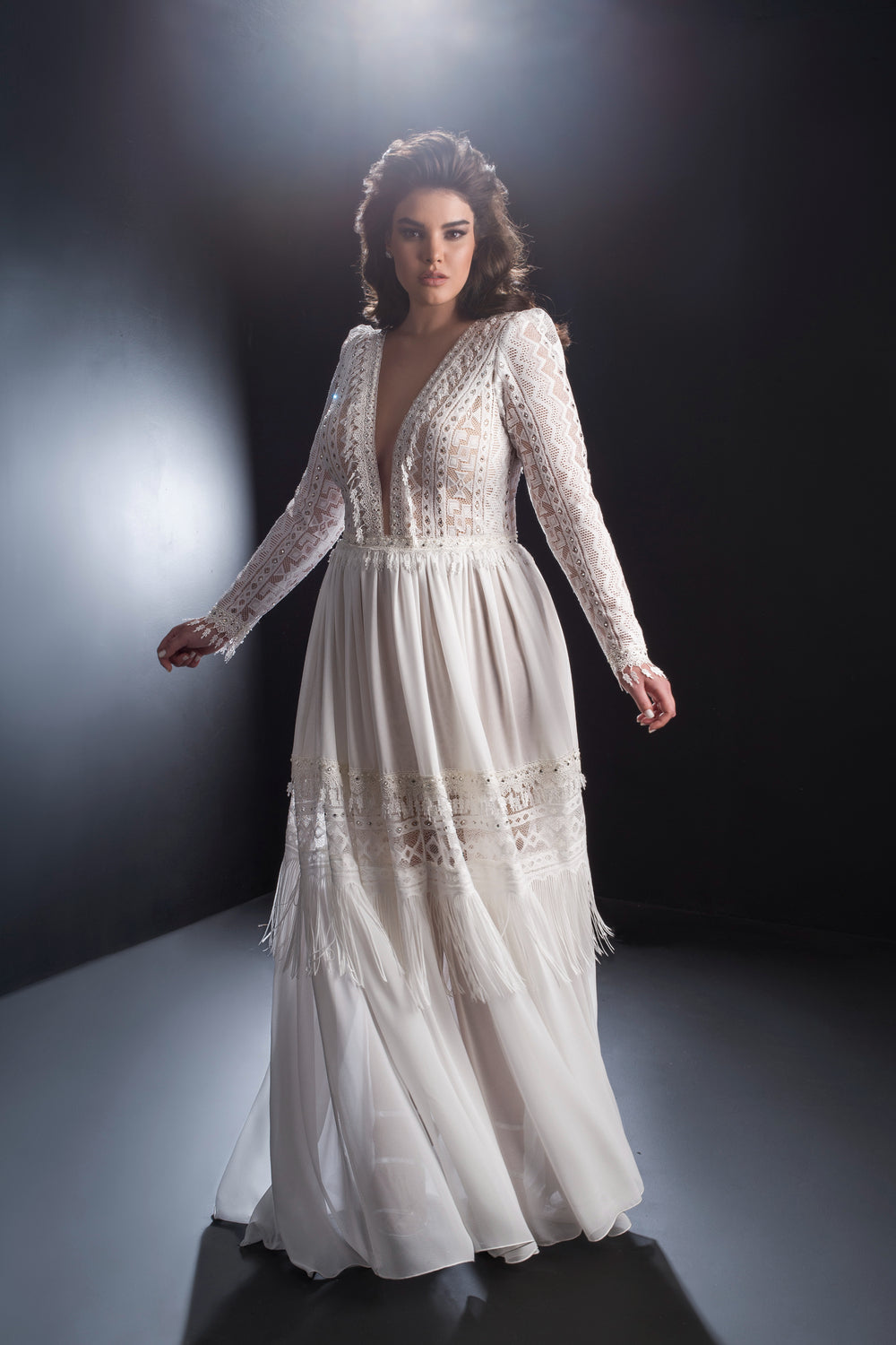 אישה מקולקציית Bridal Plus 2019 - שמלת Isabella של Dror Kontento מצטלמת בחינניות על רקע כהה. השמלה כוללת פרטים מתחרה, שרוולים ארוכים, מחשוף V עמוק ודוגמאות שקופות ועדינות.