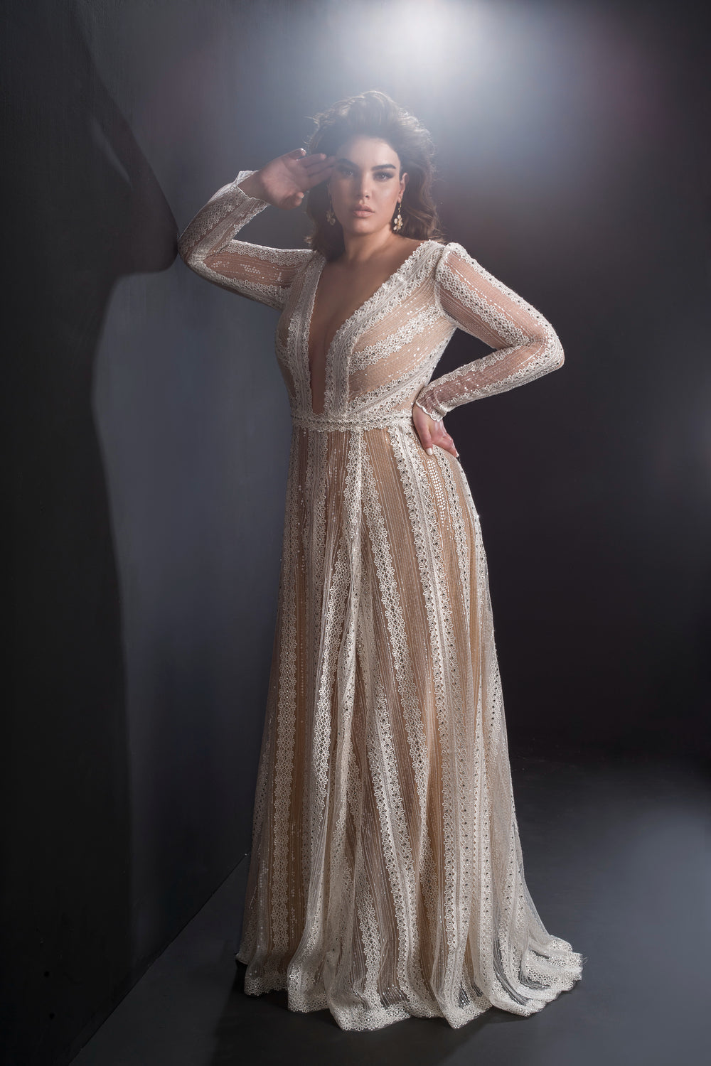 אישה מדגמנת את קולקציית Bridal Plus 2019 - Clare, שמלת תחרה ארוכה ושקופה עם מחשוף V עמוק ושרוולים ארוכים, עומדת על רקע כהה בתאורה דרמטית.