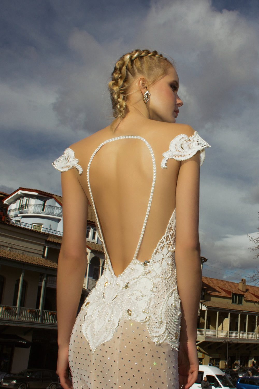 אישה בלונדינית בשמלה לבנה ללא גב עם פנינים ותחרה מקולקציית הכלות 2016 - 02 עומדת בחוץ על רקע בניינים ושמיים מעוננים - השראה מושלמת לשמלות כלה בעיצוב אישי.