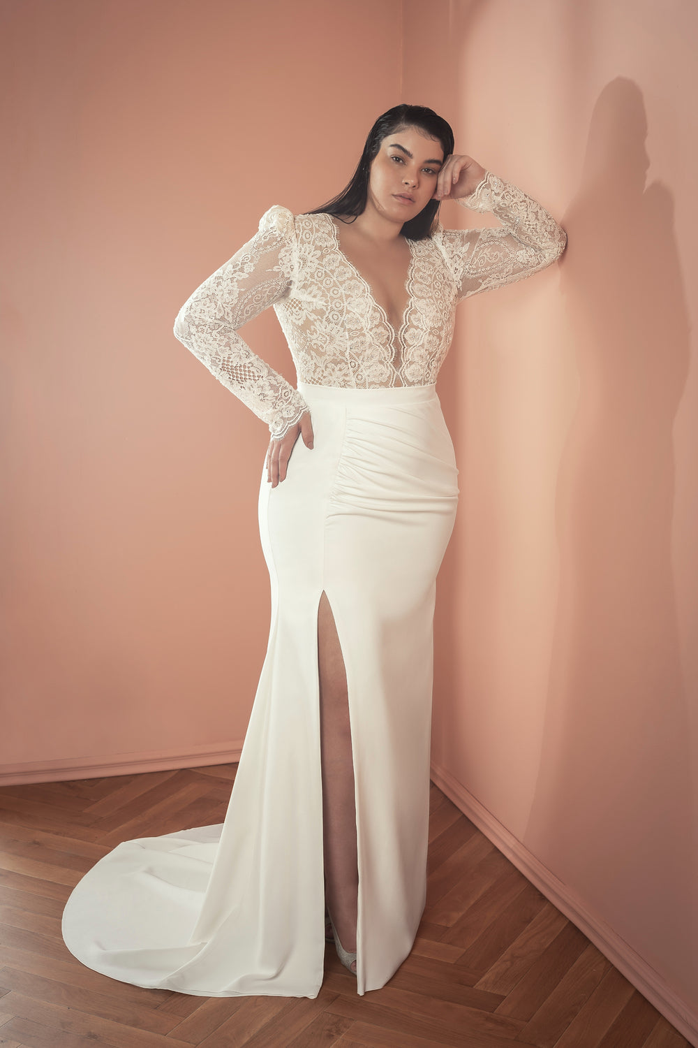 אישה מקולקציית Bridal Plus 2021 - שמלת כלה לבנה עם שרוולים ארוכים מתחרה, מחשוף V עמוק וחתך גבוה, עומדת על רקע קיר בצבע אפרסק על רצפת עץ, ומביטה בביטחון למצלמה.