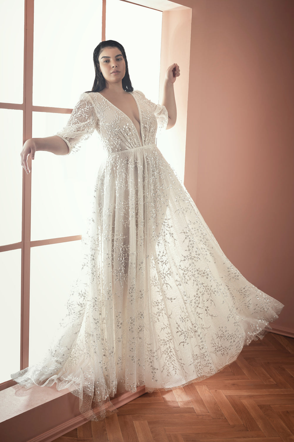 אישה בשמלת Ramona מקולקציית Bridal Plus 2021 עומדת ליד חלון גדול, ואור השמש הרך מדגיש את התחרה הלבנה והדוגמאות הפרחוניות. החדר בעל קירות בגוון אפרסק ורצפת עץ.