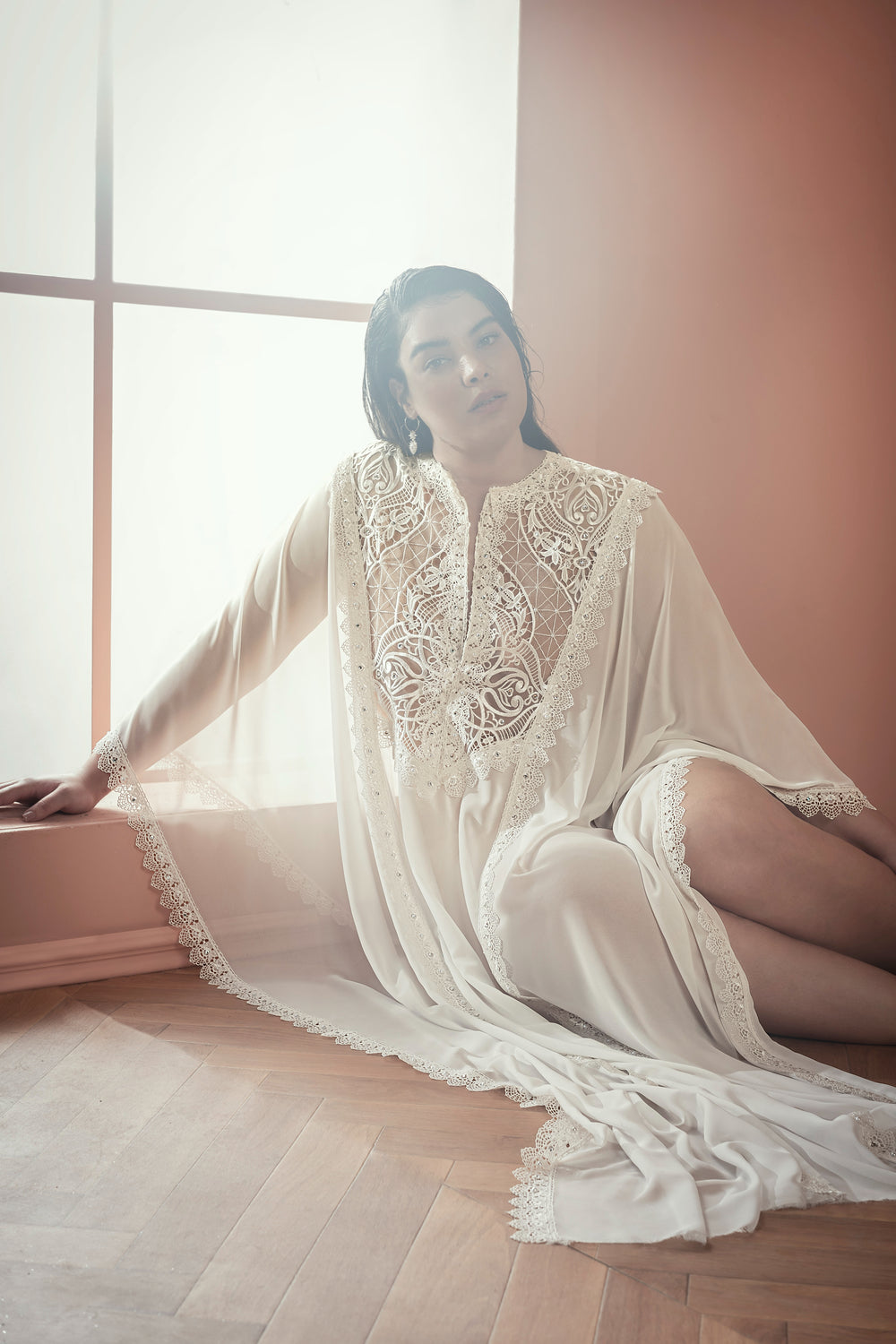 אדם לבוש בגלימה ארוכה וזורמת מתחרה לבנה "Bridal Plus Collection 2021 - Cynthia" יושב על רצפת עץ ליד חלון גדול, מביט למעלה בעוד אור טבעי רך מדגיש את פרטי התחרה העדינים של הבגד.