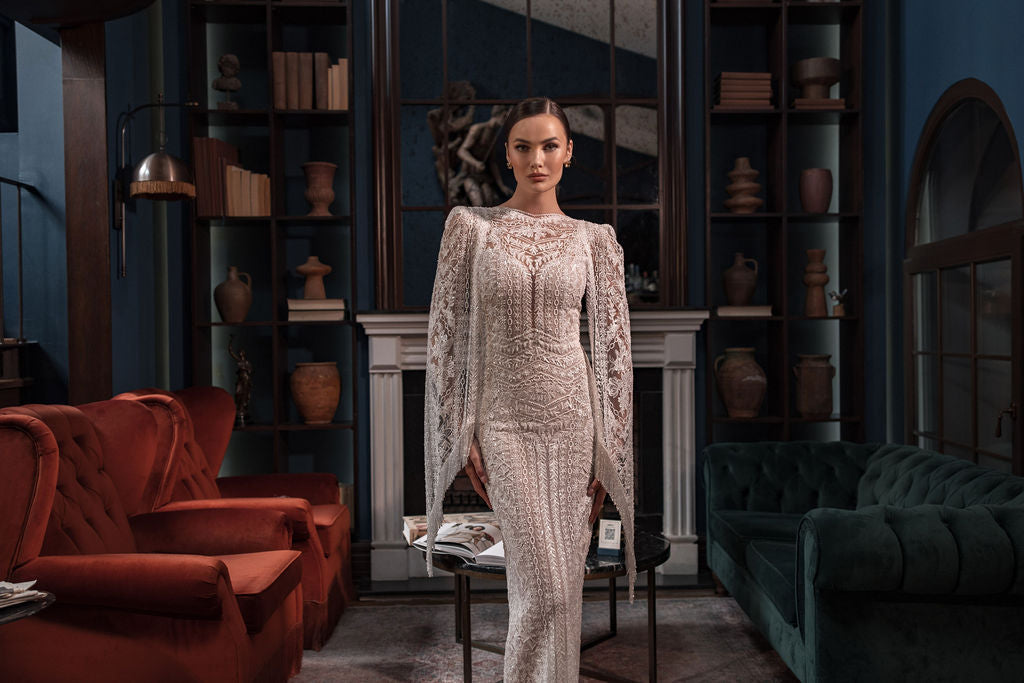 אישה בשמלת "Bridal Collection 2023 - Stormi" של דרור קונטנטו עומדת בספרייה אלגנטית, בין ספות קטיפה אדומות וירוקות עם מדפי ספרים, כלי חרס, אח ומראה המשלימים את התמונה המעודנת.